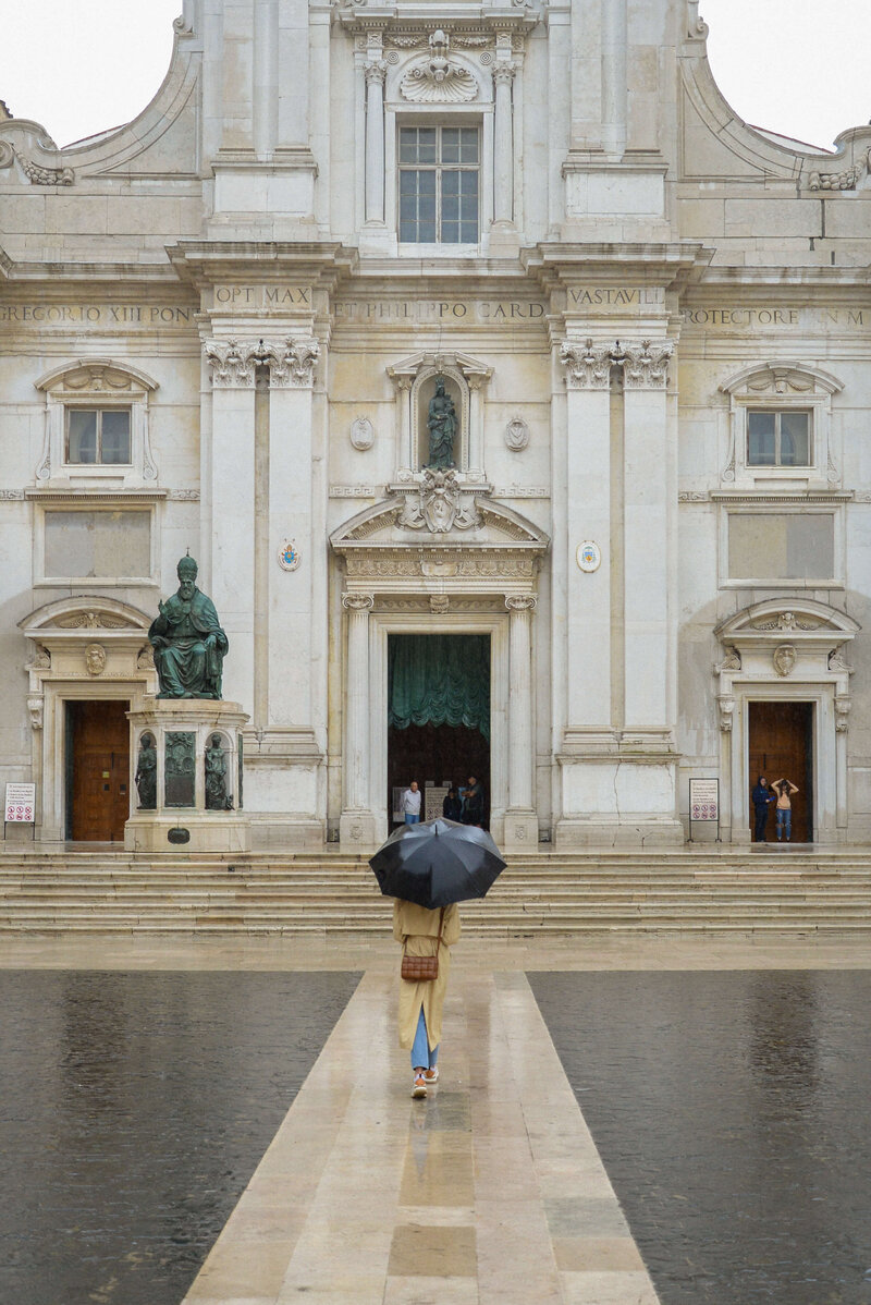 A traveler walks towards an ornate Italian church, holding an umbrella on a rainy day, capturing the beauty of exploring Italy's hidden gems.