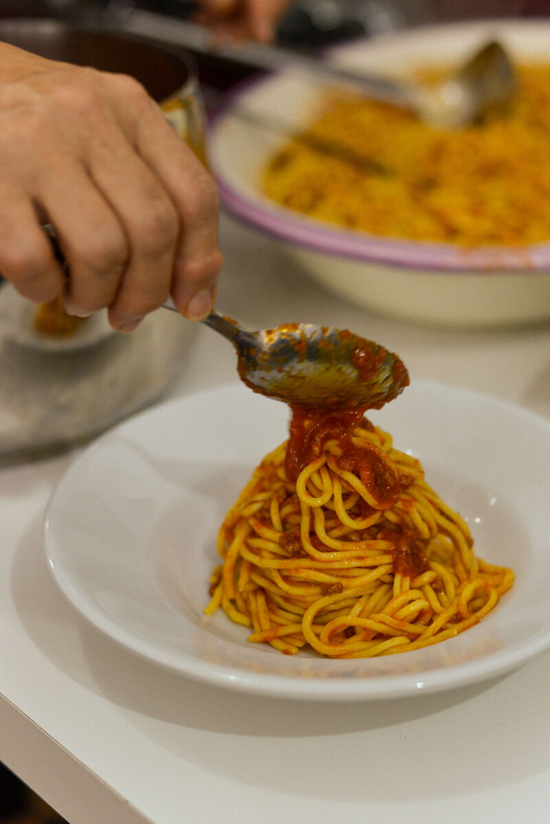 A hand serves a portion of rich, saucy spaghetti onto a plate, emphasizing the joy of gluten-free dining in Italy.
