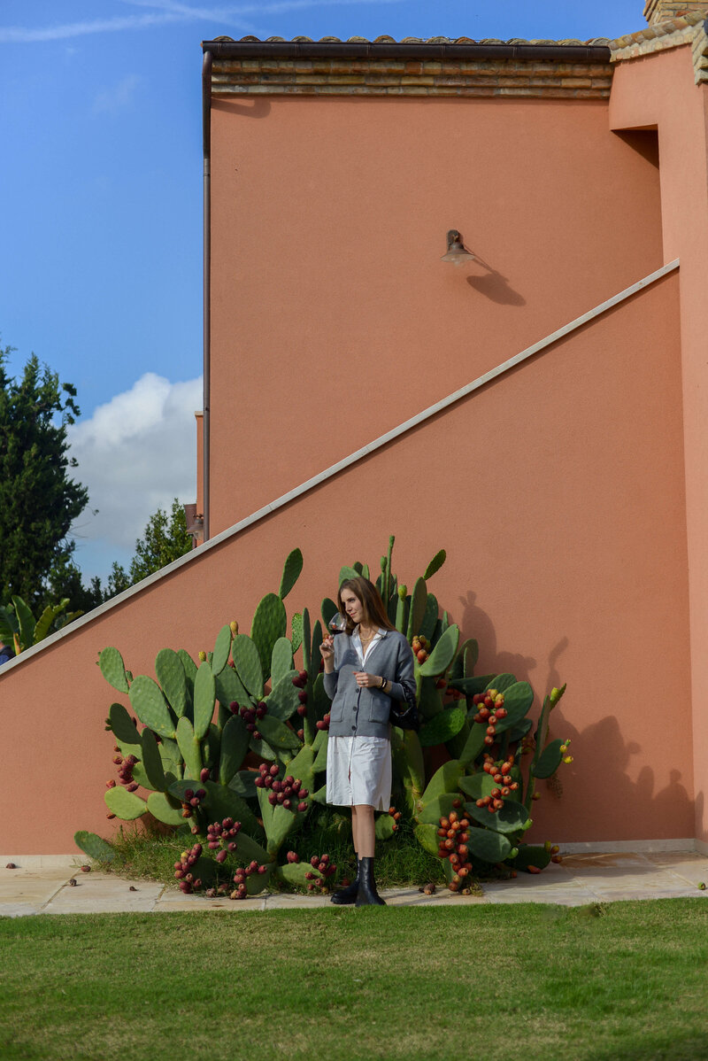 A traveler sips a drink while standing near a cactus in front of a bold orange building, embracing the charm of an authentic Italian experience.