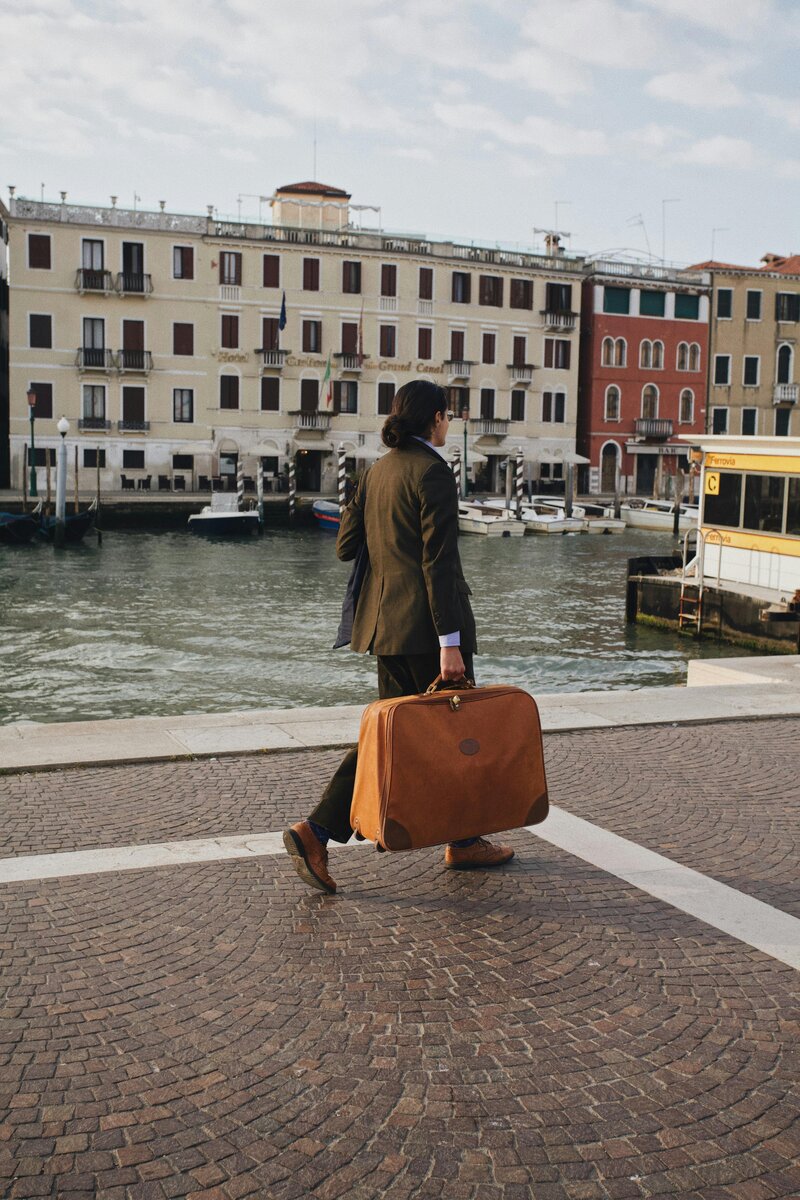 A traveler in a suit walks along a cobblestone street carrying a vintage-style suitcase, symbolizing preparation for an Italian journey.