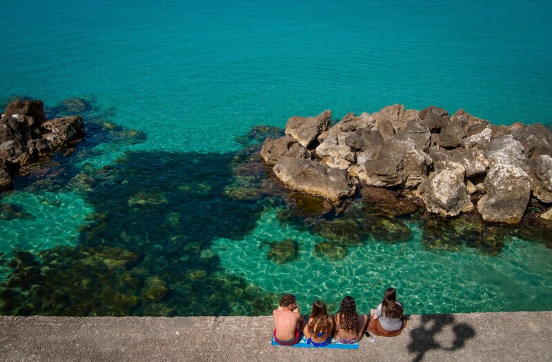 a group of people sit on a rock ledge next to the blue-green Italian ocean On the Italian on Tour’s 8 day Passport to Puglia Food & Wine Tour